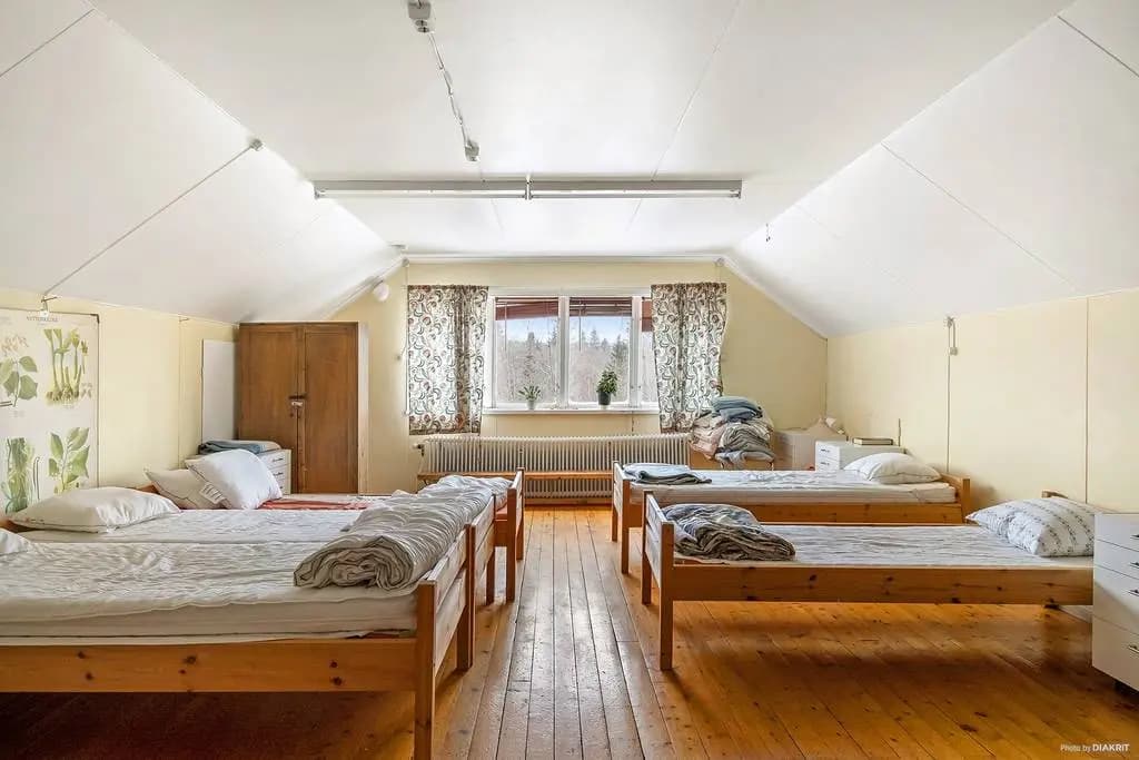 A dormitory with 6 beds lined along two slanted walls with a window overviewing the forest.